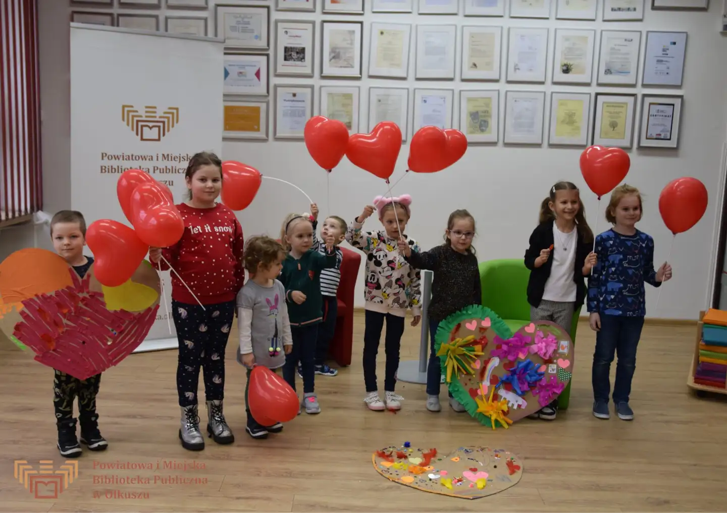 Grupa dzieci stojąca i pozująca do wspólnego zdjęcia. W rękach trzymają baloniki - czerwone serduszka. Obok nich leżą pieknie ozdobione serca z kartonów.