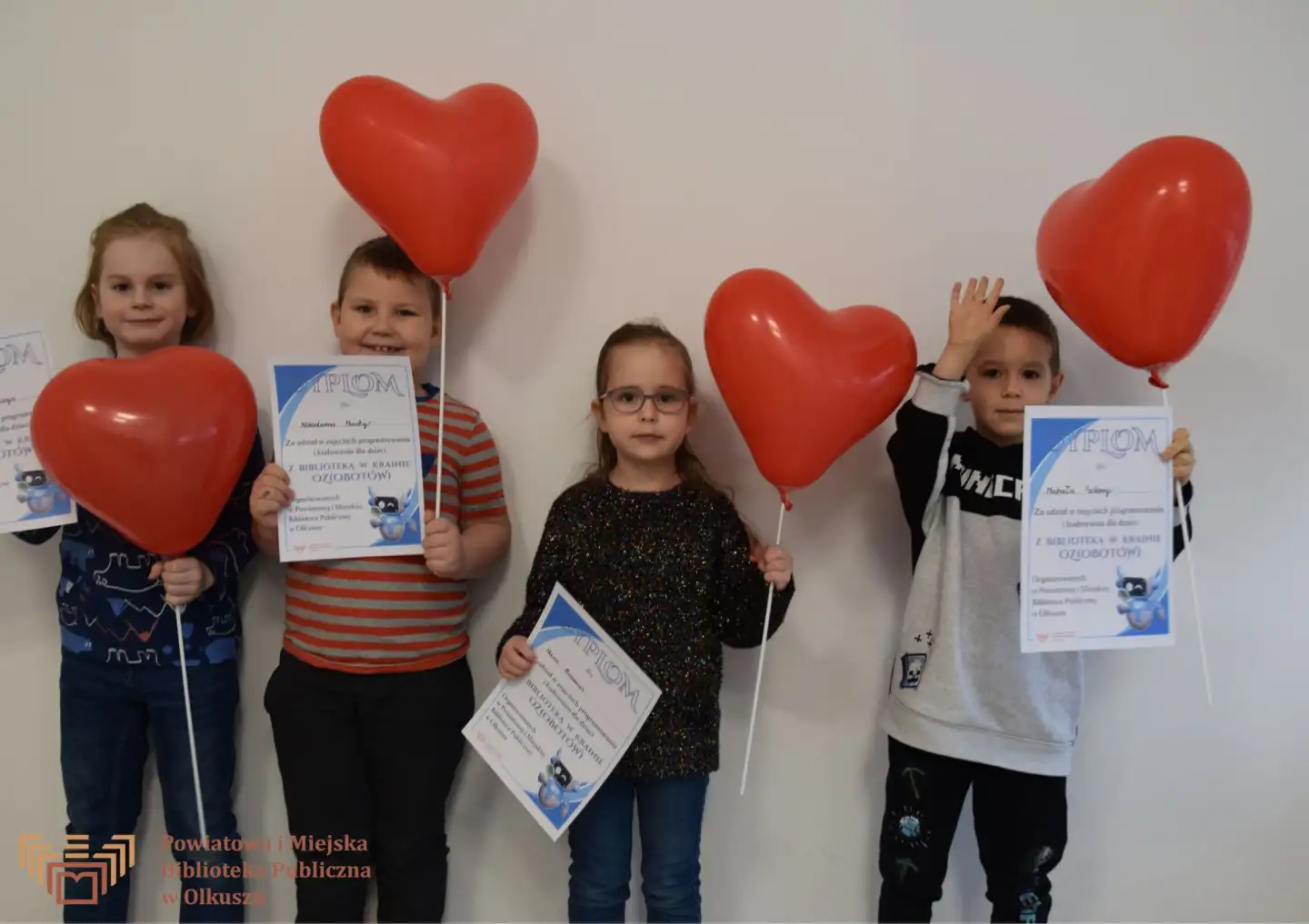 Grupa dzieci stoi przy ścianie i pozuje do wspólnego zdjęcia. W rękach trzymają balony w kształcie serca oraz dyplomy.