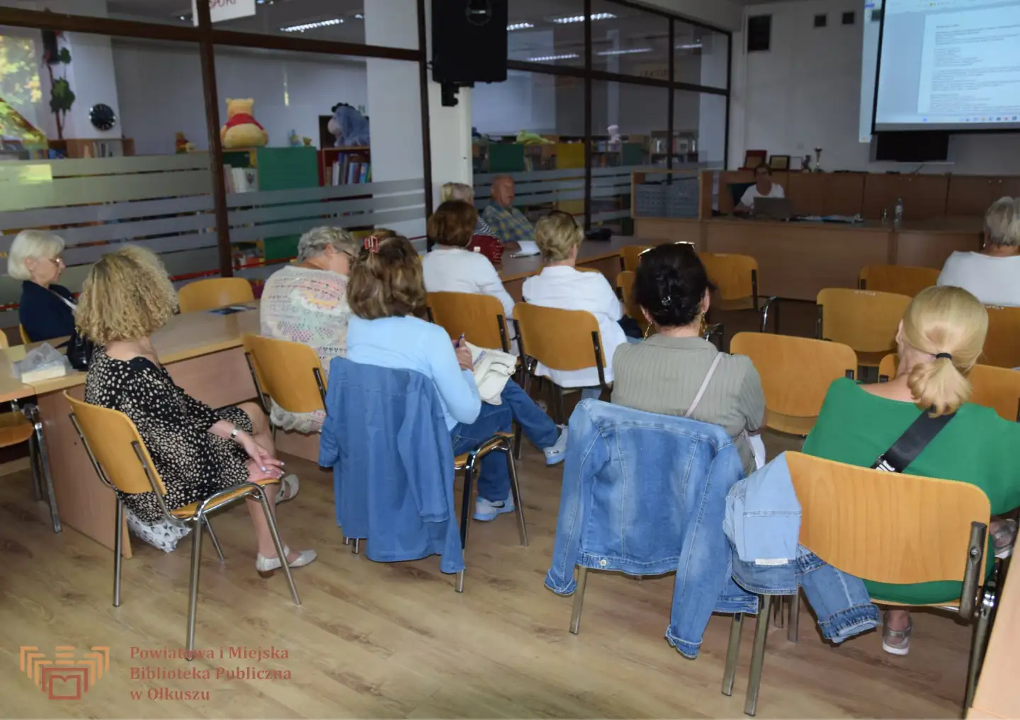 Grupa osób siedząca na krzesłach w sali konferencyjnej w trakcie prezentacji. W tle prezentacja i wypożyczalnia książek dla dzieci