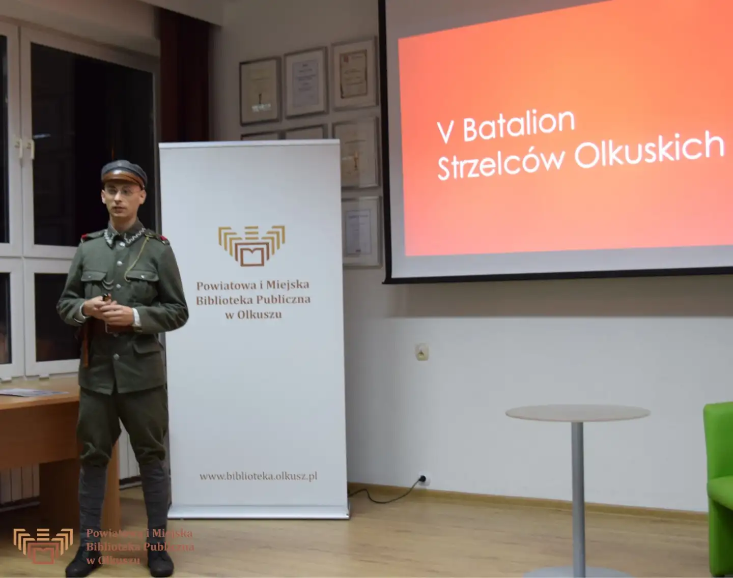 Zdjęcie przedstawia młodego mężczyznę w mundurze żołnierza AK. Za nim stoi biały baner z logo i napisem Powiatowa i Miejska Biblioteka Publiczna w Olkuszu oraz ekran z wyświetlonym slajdem i napisem: V Batalion Strzelców Olkuskich.