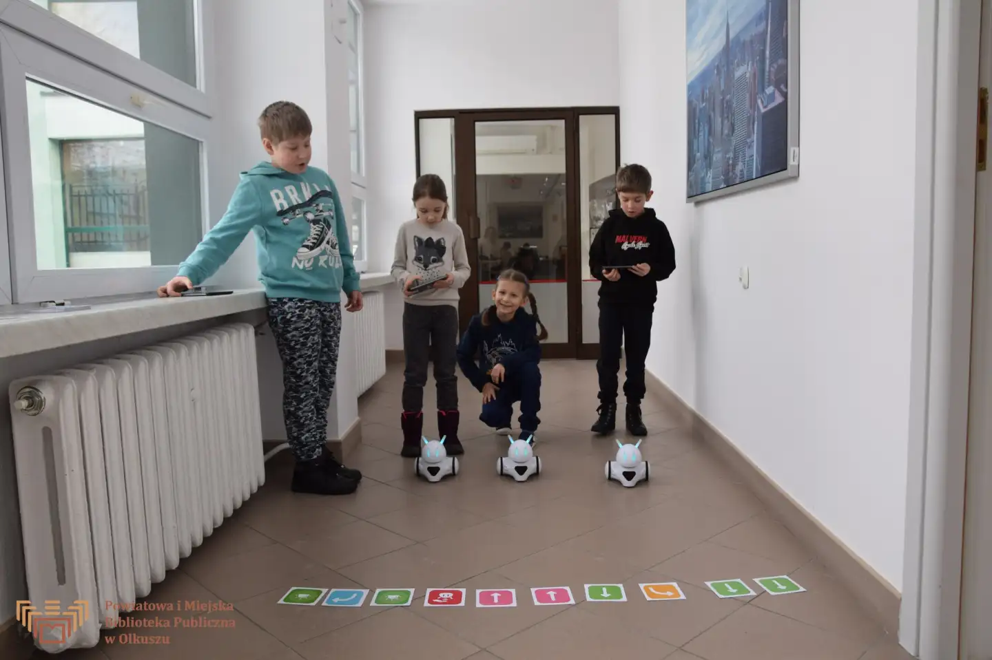 Zdjęcie przedstawia 4 dzieci stojącą na korytarzu Biblioteki. Przed dziećmi widoczne są trzy roboty (photon'y) oraz kartki ze strzałkami.