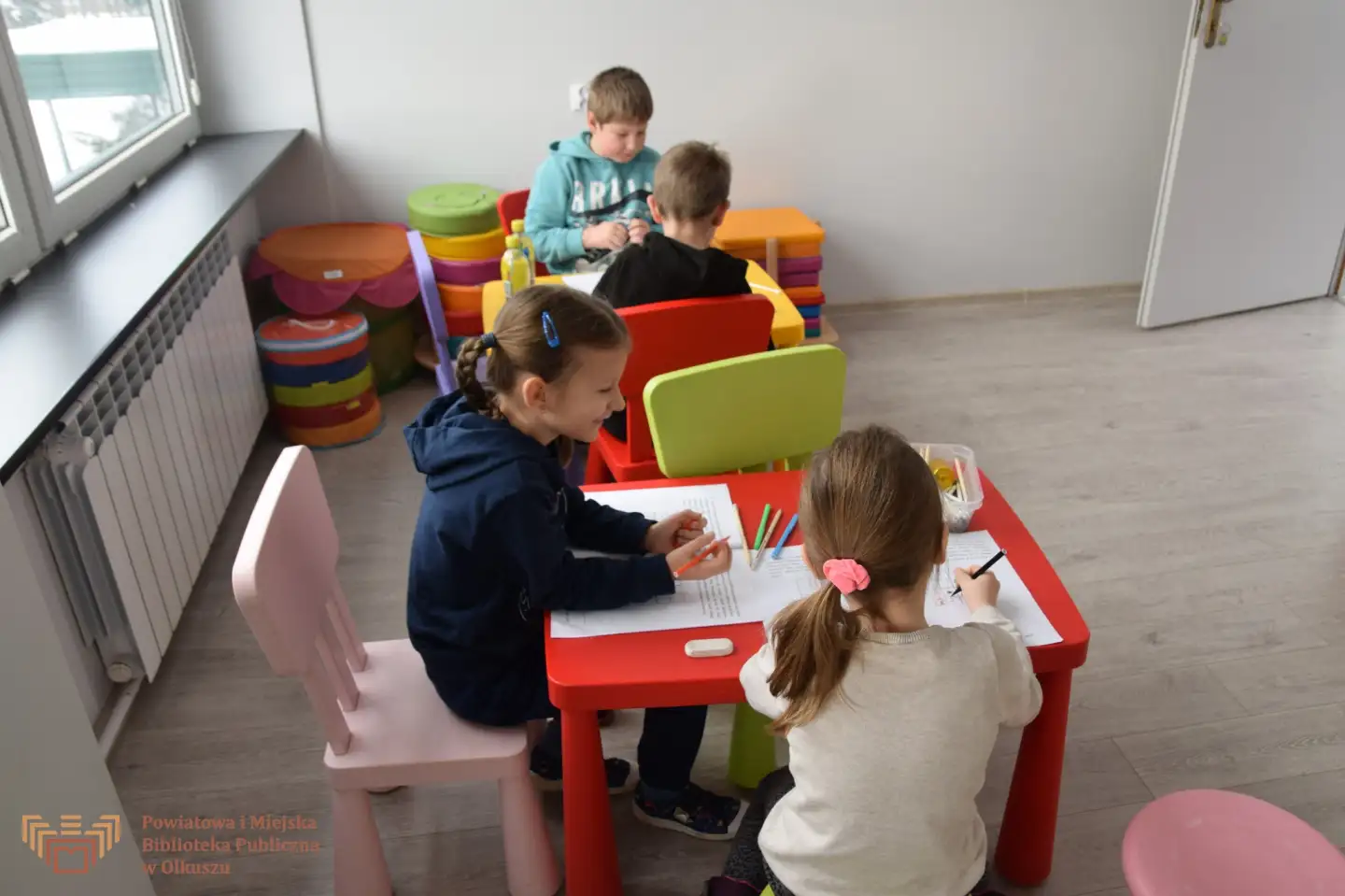 Zdjęcie zostało zrobione w sali edukacyjnej Biblioteki. Przedstawia czwórkę dzieci, które siedzą przy stoliczkach i wypełniają zadania.