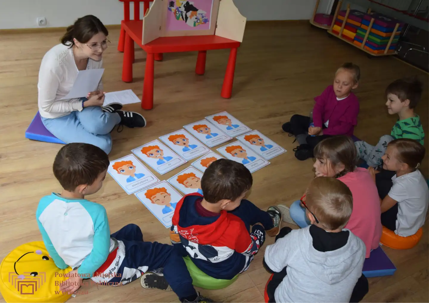Dzieci siedzące na podłodze wokół kartek przedstawiających twarz chłopca z rudymi włosami. Każdy obrazek przedstawia inną emocję. Obok siedzi Pani prowadząca, uśmiechająca się do dzieci