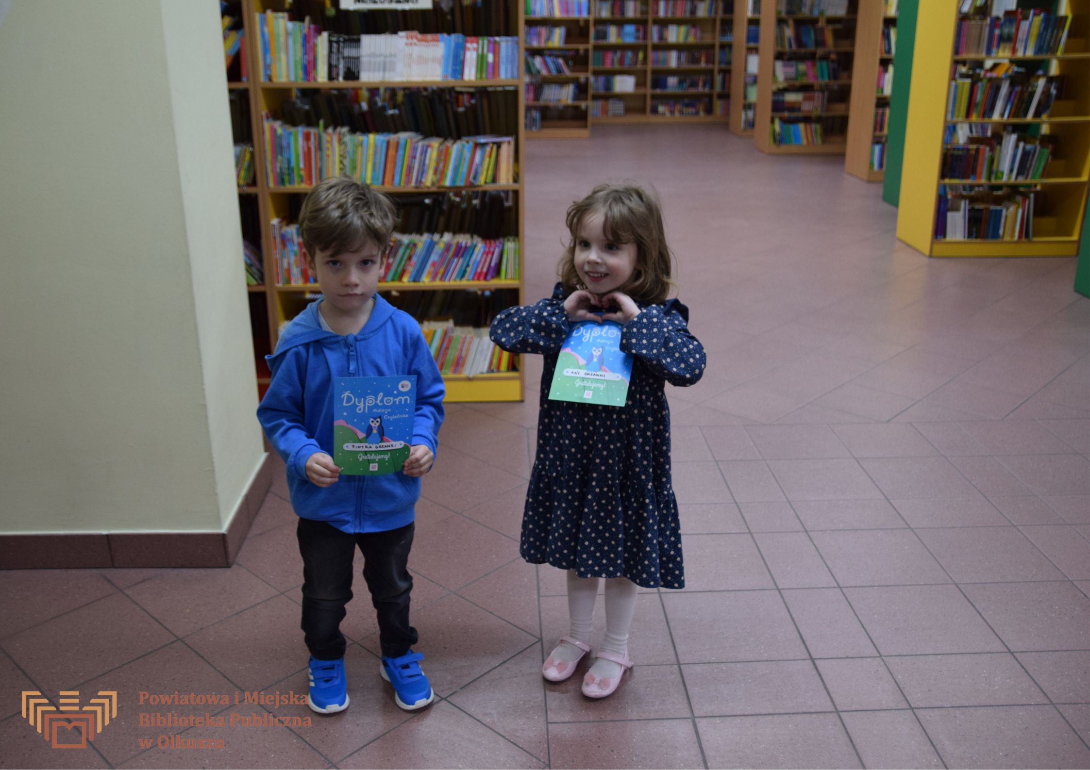 Na pierwszym planie chłopczyk i dziewczynką, trzymający dyplom akcji MAŁA KSIĄŻKA – WIELKI CZŁOWIEK, w tle regały z książkami