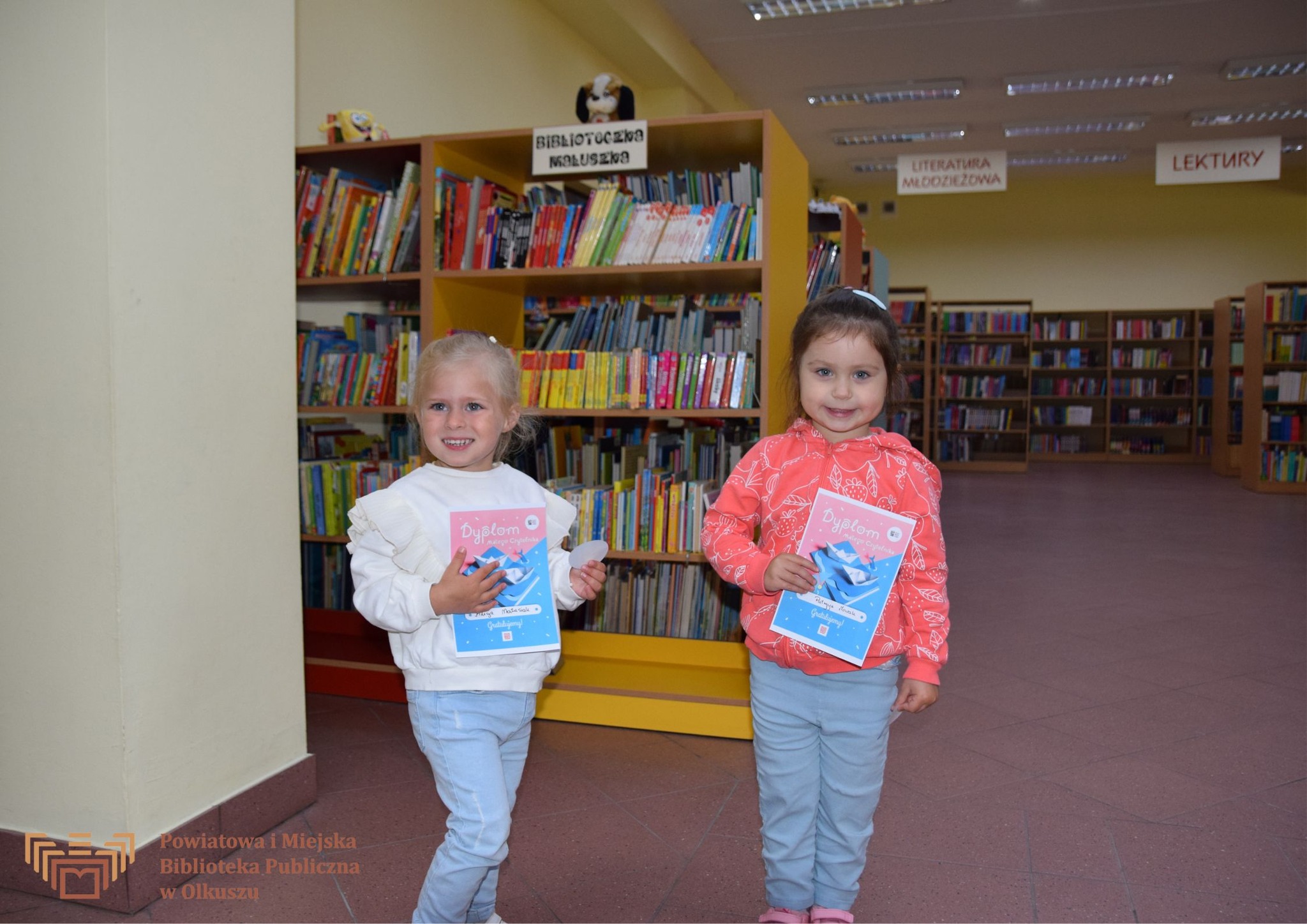 Dwie uśmiechnięte dziewczynki trzymające dyplomy, w tle regały z książkami