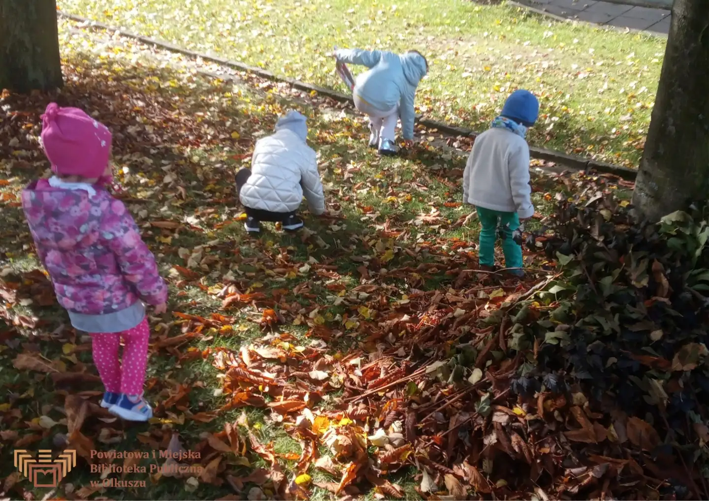 Zdjęcie zostało zrobione przed budynkiem Biblioteki. Przedstawia czwórkę dzieci zbierające liście jesienią.