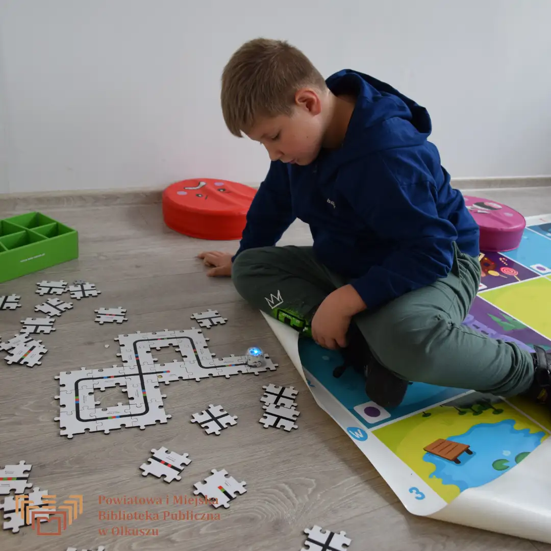 Zdjęcie przedstawia chłopca siedzącego na kolorowych poduszkach na podłodze. Układa z puzzli trasę dla małego ozobota.
