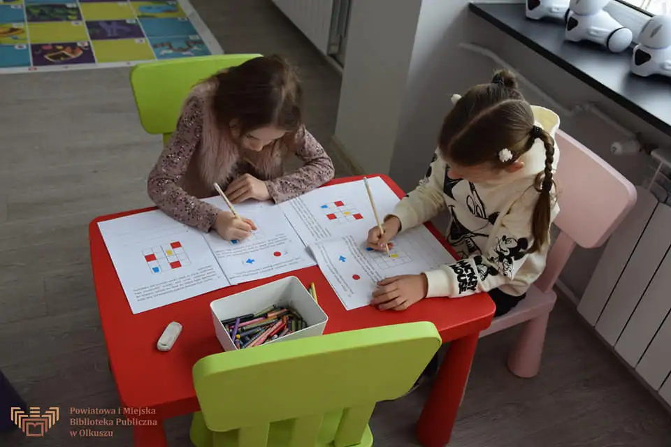 Zdjęcie przedstawia dwie dziewczynki siedzące przy czerwonym stoliku. Rozwiązują przygotowane dla nich zadania.