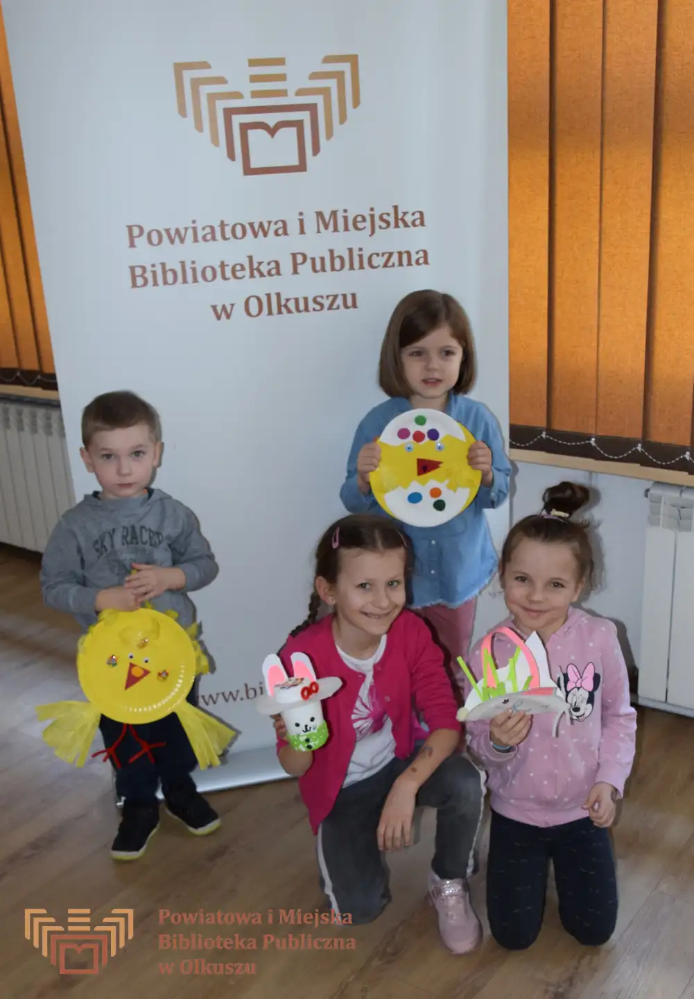 Czwórka dzieci stoi przed szyldem PiMBP w Olkuszu i pozuje do zdjęcia. Dzieci trzymają samodzielnie zrobione prace plastyczne nawiązujące do Wielkanocy.