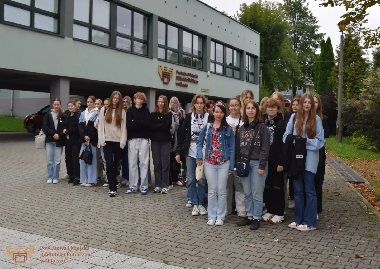 Zdjęcie zostało zrobione przed budynkiem olkuskiej Biblioteki. Przedstawia dużą grupę młodzieży licealnej.