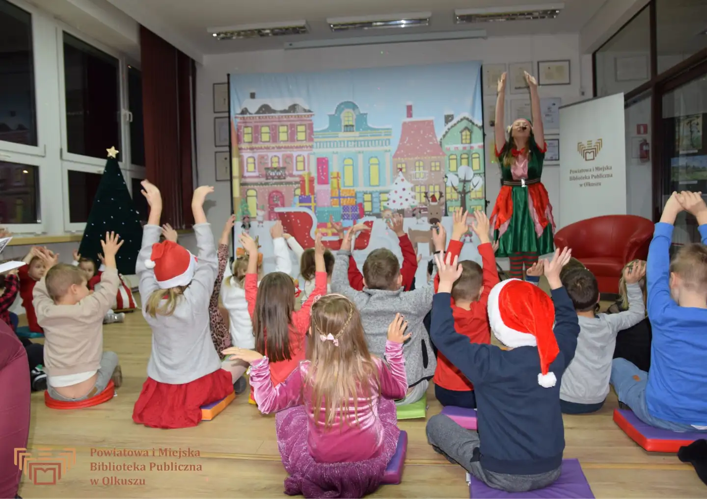 Zdjęcie zostało zrobione w Czytelni olkuskiej Biblioteki. Na tle płótna z zimowy, świątecznym miastem stoi kobieta w stroju elfa z uniesionymi do góry rękoma. Na podłodze siedzą dzieci i również podnoszą ręce do góry. Niektóre z nich mają na głowach czapki Mikołajowe.