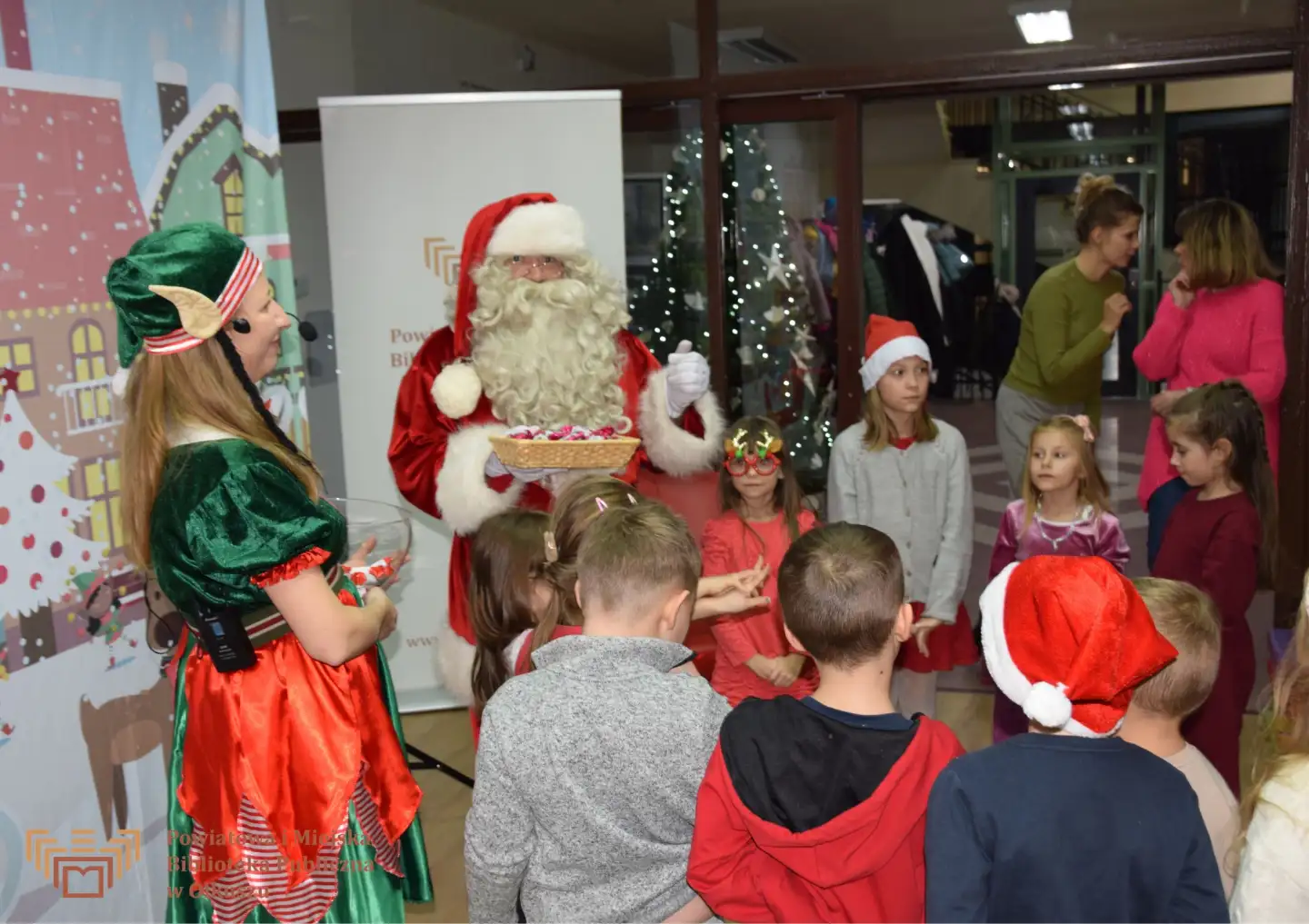 Zdjęcie zostało zrobione w Czytelni olkuskiej Biblioteki. Na tle płótna z zimowy, świątecznym miastem stoi Mikołaj, kobieta w stroju elfa i ustawione w kółko dzieci. Mikołaj i elf trzymają rękach miski ze słodkościami dla dzieci.