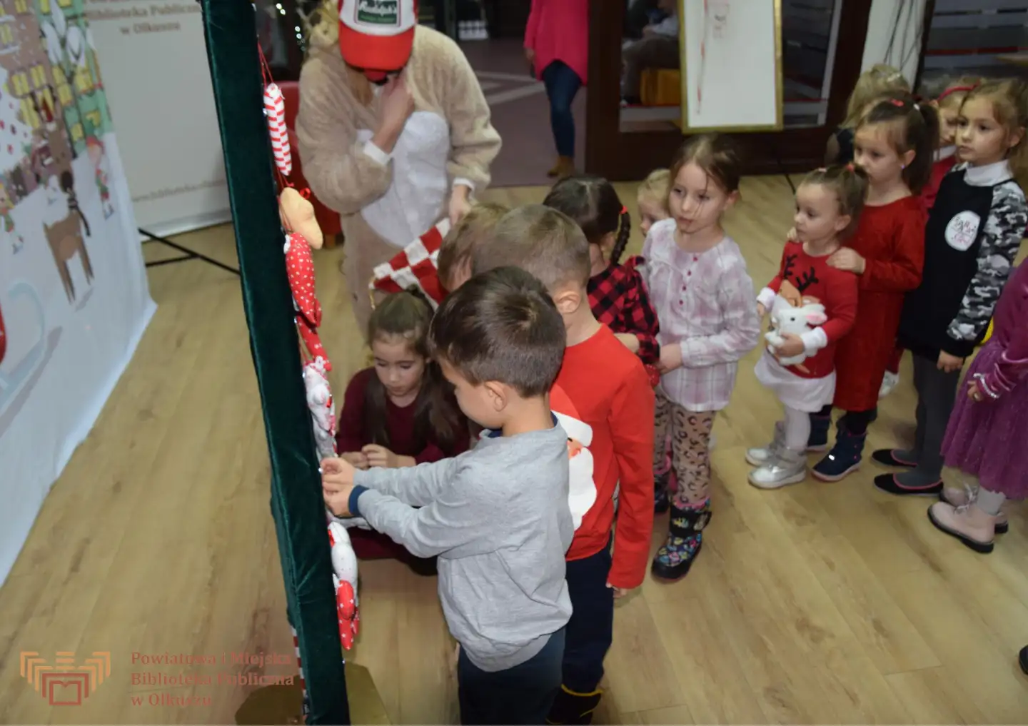 Zdjęcie zostało zrobione w Czytelni olkuskiej Biblioteki. Do tablicy w kształcie choinki stoją ustawione dzieci i przypinają ozdoby świąteczne.