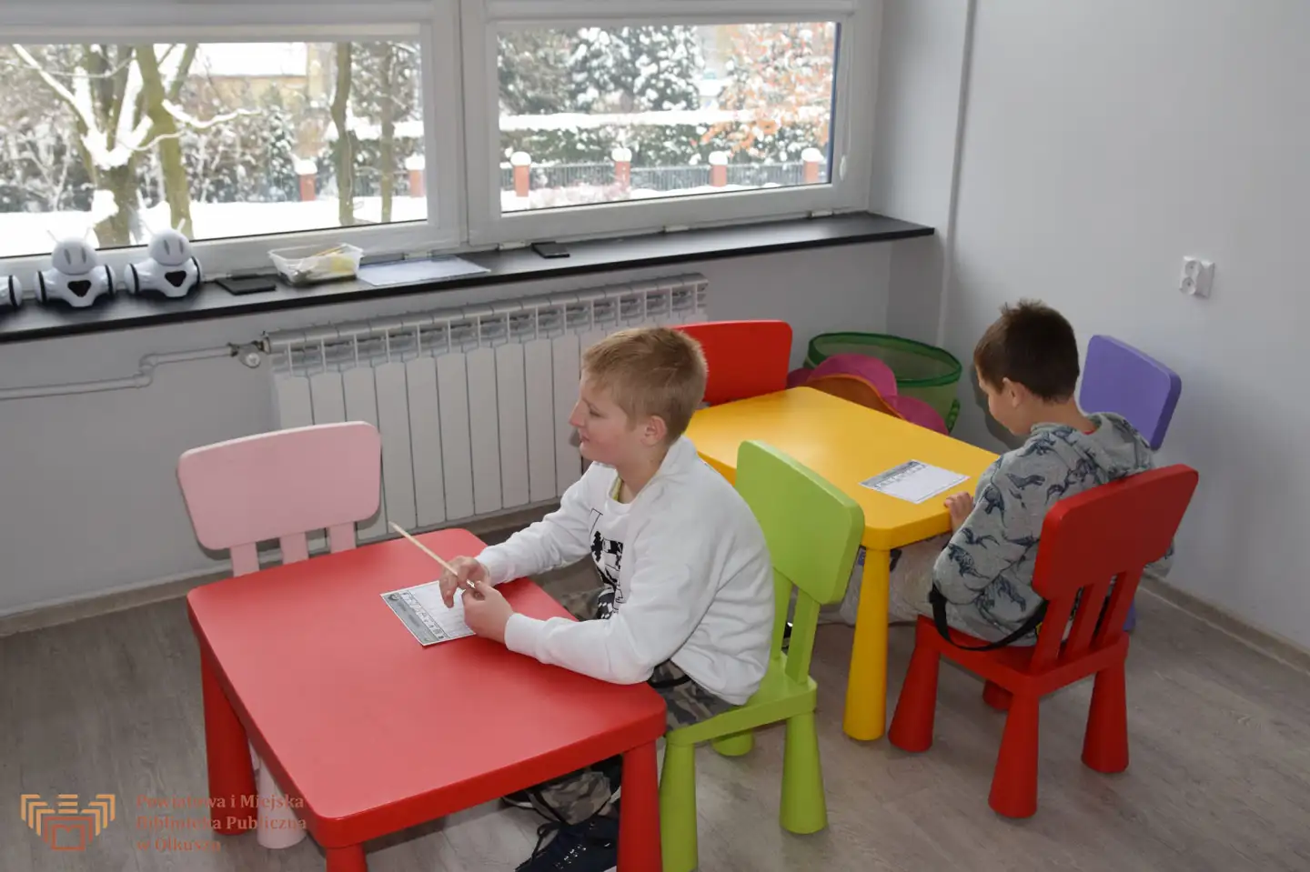 Zdjęcie przedstawia dwóch chłopców siedzących przy kolorowych stolikach. Rozwiązują przygotowane dla nich zadania.