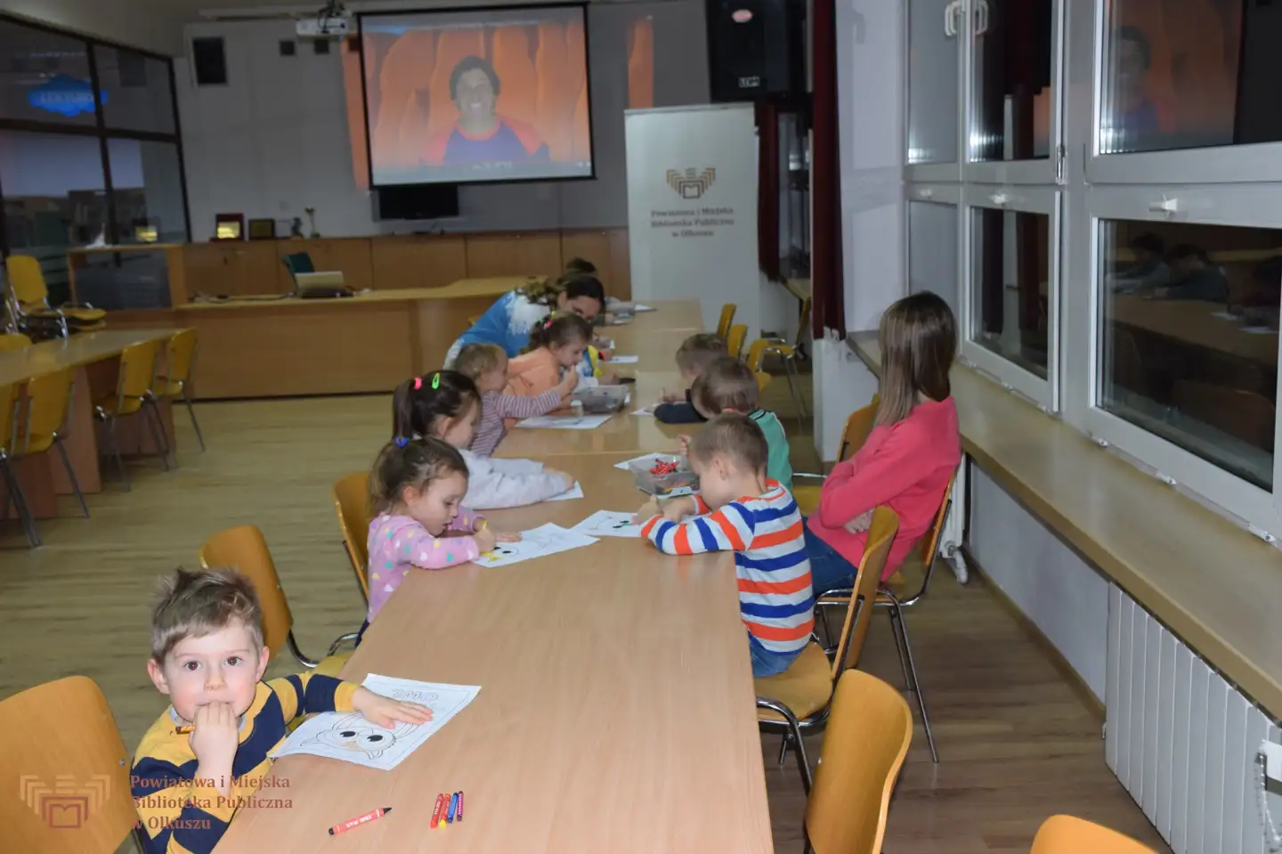 Zdjęcie zostało zrobione w czytelni Biblioteki. Przedstawia grupę dzieci siedzących przy stolikach i malujących obrazek sowy.