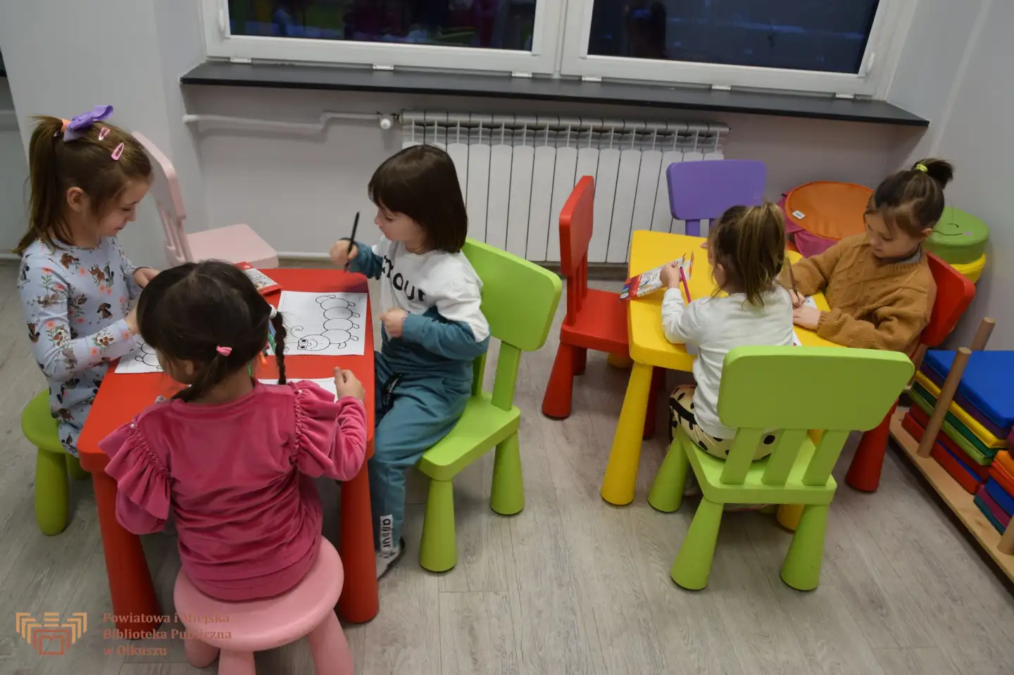 Zdjęcie zostało zrobione w sali edukacyjnej olkuskiej Biblioteki. Przedstawia dzieci siedzące przy kolorowych stolikach i malujących kolorowymi kredkami gąsienice.