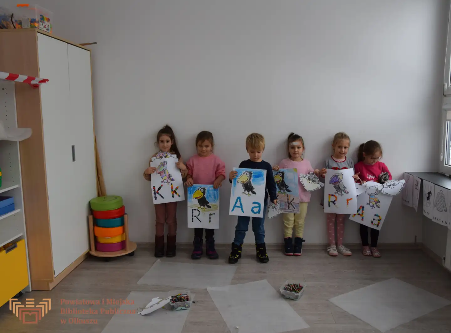 Zdjęcie przedstawia grupę dzieci, które pozującą do zdjęcia. Dzieci stoją pod białą ścianą i trzymają duże kartki. Na kartkach przedstawione są papugi i litery K, A, R wyklejone plasteliną.