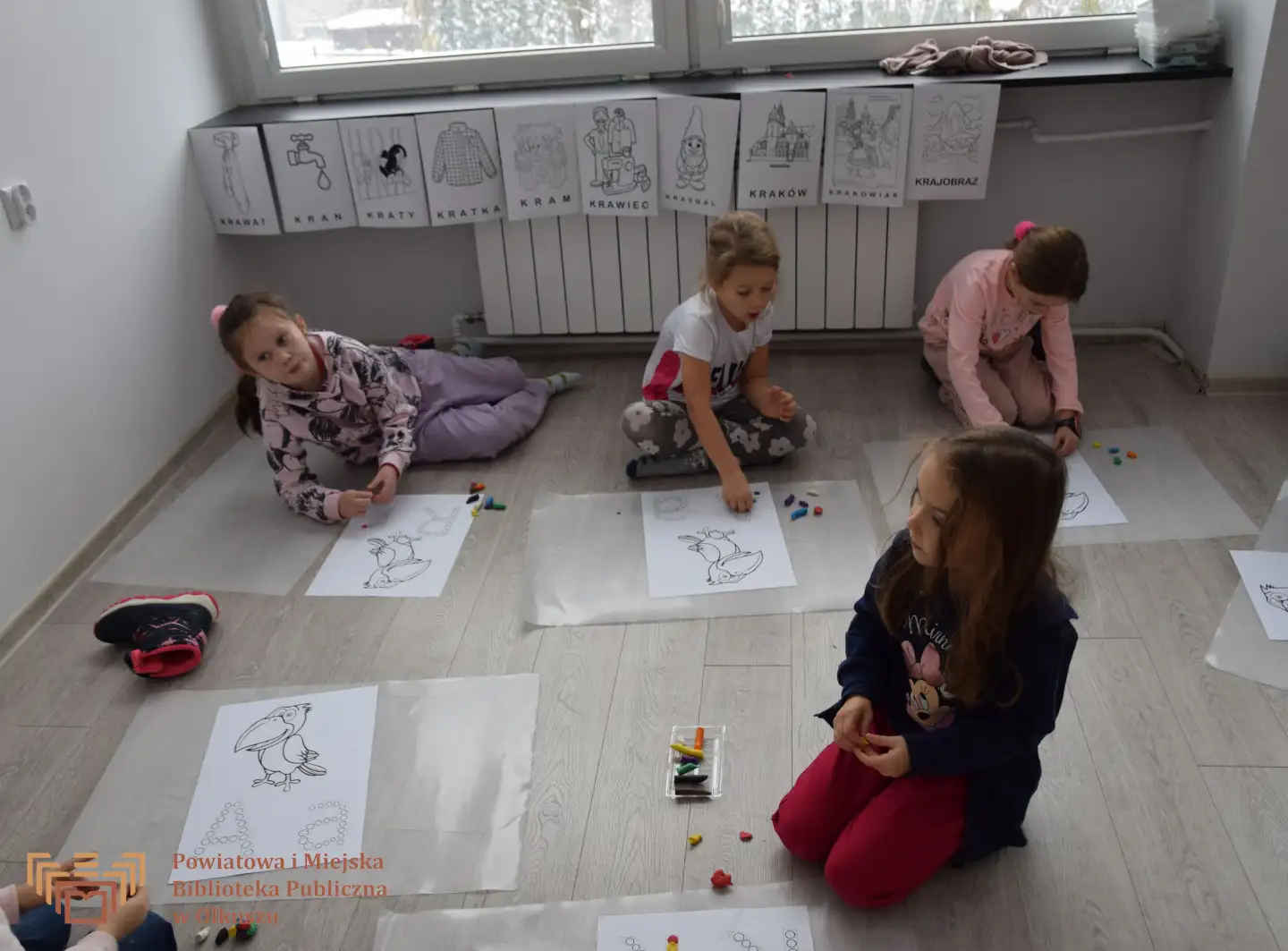 Zdjęcie przedstawia grupę dzieci, które siedzą na podłodze. Przed dziećmi znajdują się kartki z kolorowanką w kształcie papugi oraz liter K, A. Dzieci przyklejają plastelinę do obrazków.