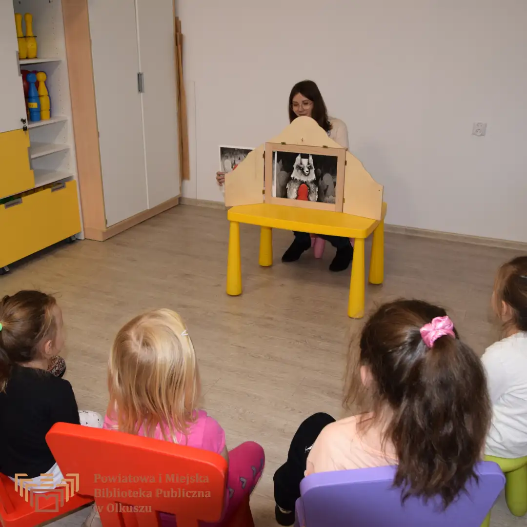 Zdjęcie przedstawia dzieci siedzące na kolorowych stołeczkach. Są zwrócone w stronę Pani, która siedzi przy żółtym stoliku i pokazuje dzieciom teatrzyk.