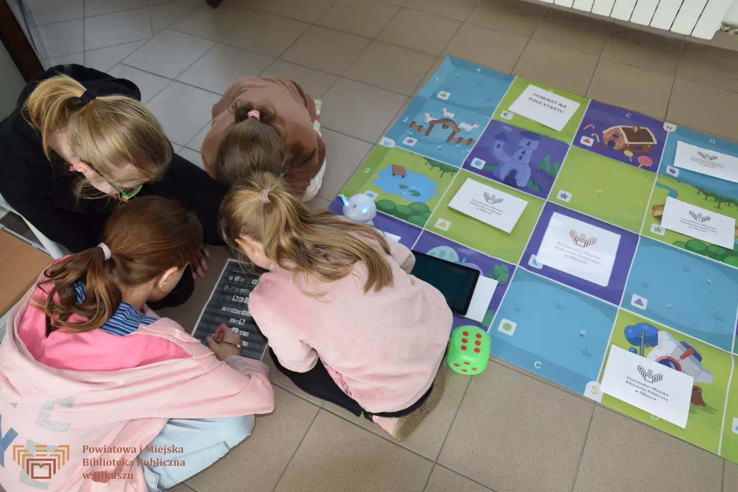 Grupa dziewczynek siedzi na podłodze i rozwiązuje zadania na kartce. Obok grupy widoczna gra planszowa w formie maty oraz robot Photon.