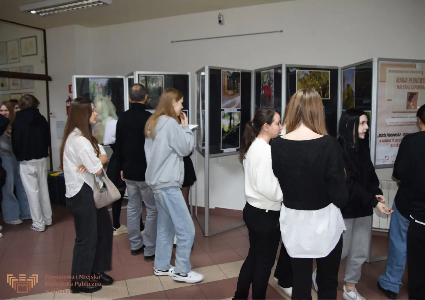 Zdjęcie przedstawia grupę młodzieży, która ogląda wystawę prac znajdującą się w holu olkuskiej Biblioteki.